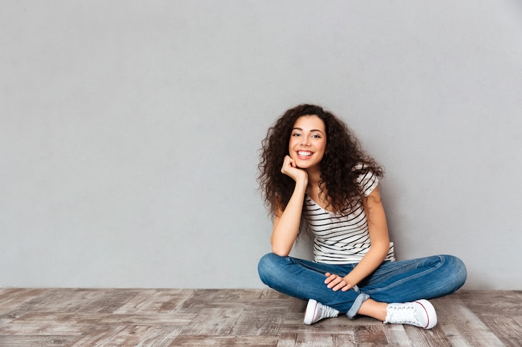 Foto grátis linda mulher encaracolada em roupas casuais, sentado em posição de lótus no chão, sustentando a cabeça com a mão, sendo feliz e sincero sobre parede cinza