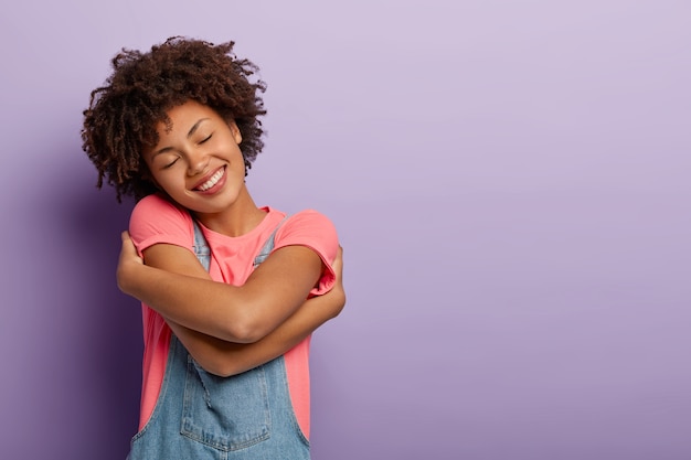 Linda mulher encaracolada se abraça de prazer, sente conforto, calma e amor, inclina a cabeça e sorri positivamente, com os olhos fechados, posa sobre a parede roxa, espaço em branco