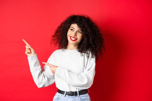 Linda mulher europeia com cabelo encaracolado e maquiagem, apontando o dedo para a esquerda e olhando para o lado com um sorriso sonhador, verificando o negócio da promoção, plano de fundo vermelho.