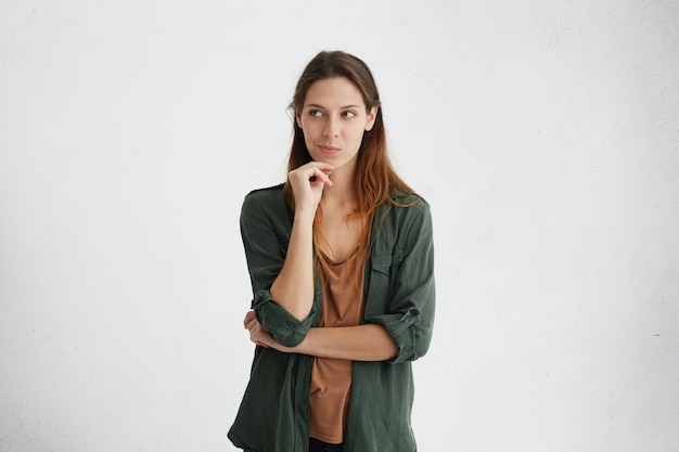 Foto grátis linda mulher europeia com longos cabelos escuros, segurando a mão no queixo, olhando pensativamente de lado, tendo uma ideia em sua mente.