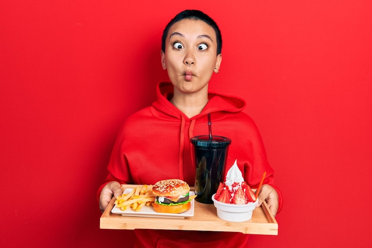 Foto grátis linda mulher hispânica com cabelo curto comendo um saboroso hambúrguer clássico com batatas fritas e refrigerante fazendo cara de peixe com a boca e olhos semicerrados loucos e cômicos