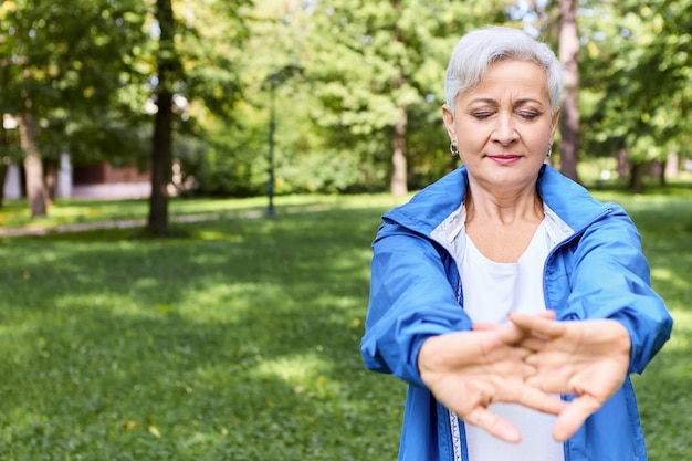 Linda mulher idosa de cabelos grisalhos com uma jaqueta de cola posando ao ar livre com os olhos fechados, esticando as mãos, fazendo exercícios de aquecimento antes do treino cardiovascular, copyspace para suas informações de publicidade