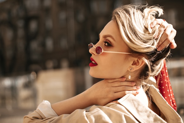 Linda mulher loira em óculos de sol vermelhos toca o cabelo. Senhora elegante com um sobretudo bege moderno a posar ao ar livre