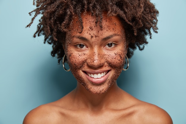 Linda mulher mestiça tem esfoliante de pele no rosto, sorri gentilmente, faz máscaras cosméticas de café, tem cabelo cacheado, ombros nus, isolado sobre parede azul