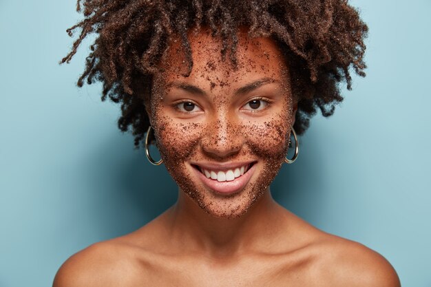 Linda mulher mestiça tem esfoliante de pele no rosto, sorri gentilmente, faz máscaras cosméticas de café, tem cabelo cacheado, ombros nus, isolado sobre parede azul