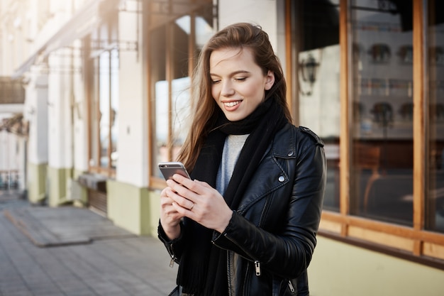 Linda mulher moderna em roupas da moda, segurando o smartphone e olhando para a tela enquanto mensagens ou navegando na net, andando na rua