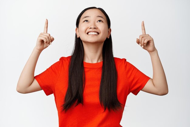 Linda mulher morena apontando os dedos para cima e sorrindo olhando para o anúncio em camiseta vermelha contra fundo branco
