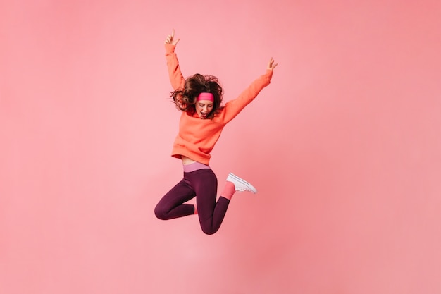 Foto grátis linda mulher morena com uma faixa esportiva rosa e um agasalho esportivo salta na parede rosa isolada