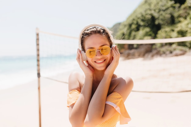 Linda mulher morena, curtindo a música com os olhos fechados na praia. foto ao ar livre de garota cativante em fones de ouvido brancos relaxando na praia selvagem.