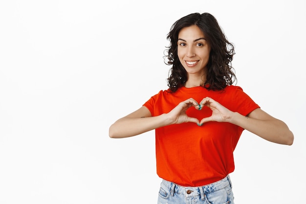 Linda mulher morena mostra o sinal do coração, sorrindo e parecendo comovente, expressa amor e ternura, gosta de você no branco