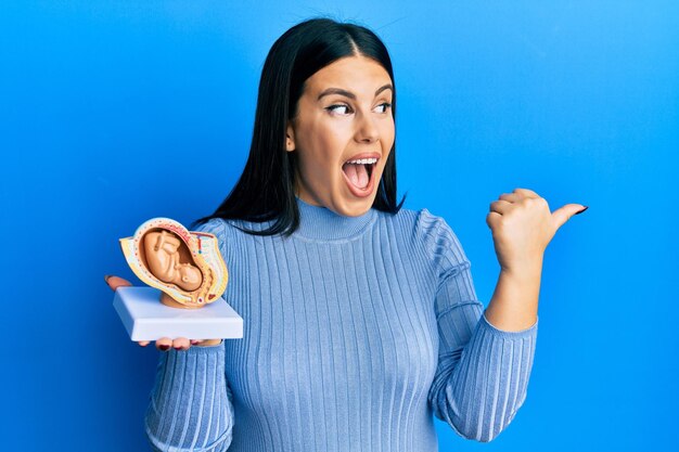 Linda mulher morena segurando o modelo anatômico do útero feminino com o feto apontando o polegar para o lado sorrindo feliz com a boca aberta