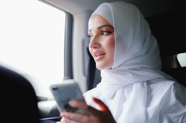 Linda mulher muçulmana usando um hijab branco, sentada no banco de trás de um carro e usando o smartphone