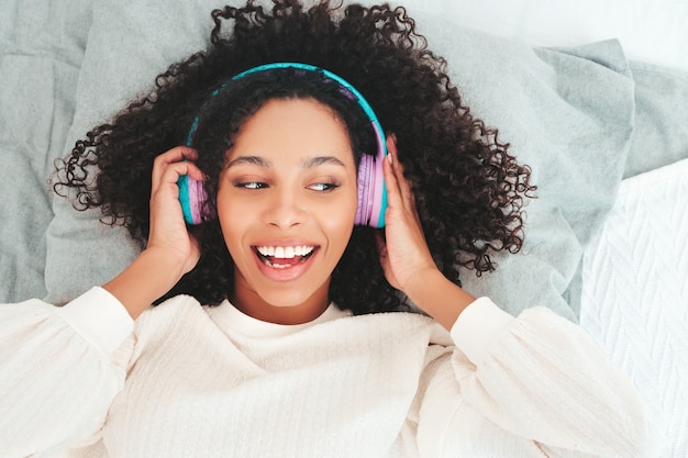 Linda mulher negra com penteado de cachos afro. Modelo sorridente de suéter e jeans. música feminina despreocupada em fones de ouvido sem fio pela manhã