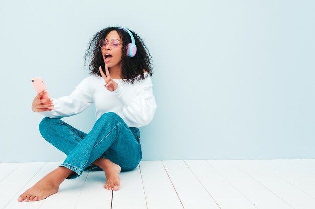 Linda mulher negra com penteado de cachos afro. Modelo sorridente de suéter e jeans. música feminina despreocupada em fones de ouvido sem fio