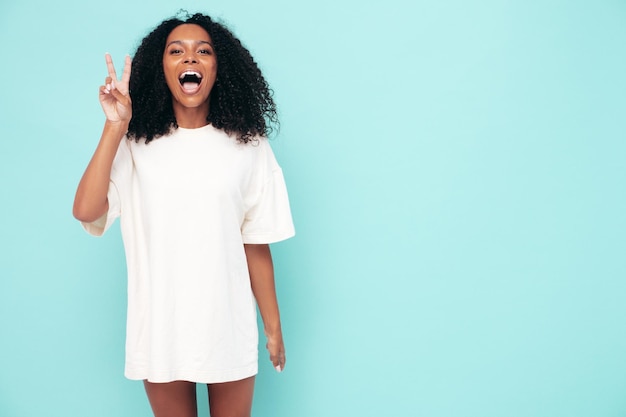 Linda mulher negra com penteado de cachos afro modelo sorridente em roupas de camiseta longa mulher despreocupada sexy posando perto da parede azul no estúdio bronzeado e alegre