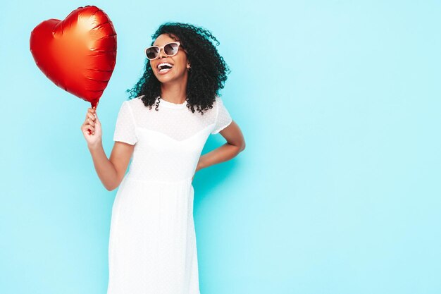 Linda mulher negra com penteado de cachos afro Modelo sorridente vestido com vestido branco de verão Mulher despreocupada sexy posando perto da parede azul no estúdio Bronzeado e alegre Segurando balão de ar de coração