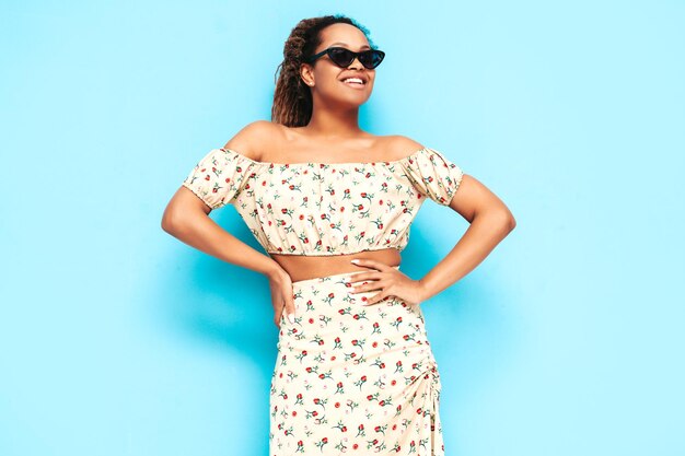 Linda mulher negra com penteado de cachos afro modelo sorridente vestido com vestido de verão mulher despreocupada sexy posando perto da parede azul no estúdio bronzeado e alegre