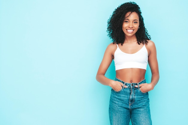 Foto grátis linda mulher negra com penteado de cachos afro modelo sorridente vestido de top branco e jeans mulher despreocupada sexy posando perto da parede azul no estúdio bronzeado e alegre isolado