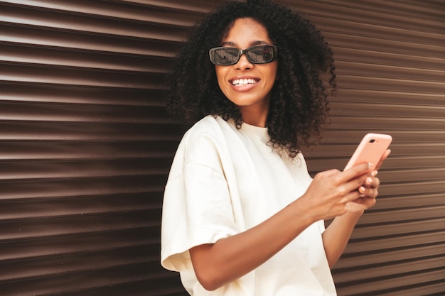 Foto grátis linda mulher negra com penteado de cachos afromodelo de hipster sorridente em camiseta branca mulher despreocupada sexy posando na rua perto de parede marrom em óculos de sololhando para a tela do smartphone usando aplicativos