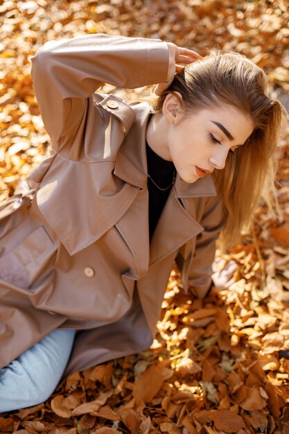 Linda mulher posando para uma foto no parque outono. Jovem sentada em folhas amarelas. Mulher loira vestindo casaco bege.