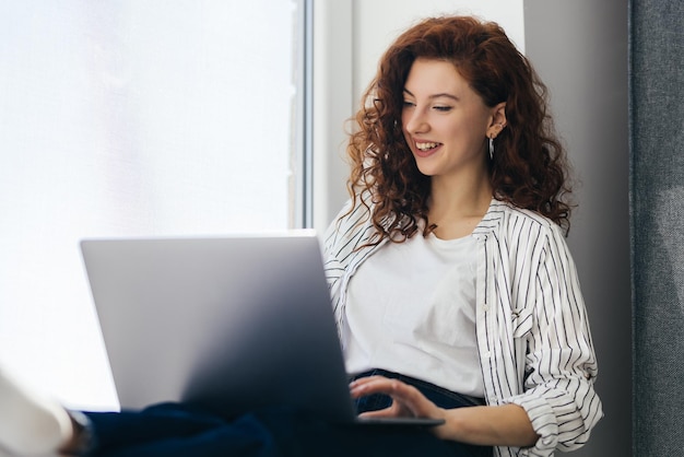 Linda mulher positiva usando laptop sentado no parapeito da janela no apartamento da cidade Jovem ruiva trabalhando em casa Conceito freelance