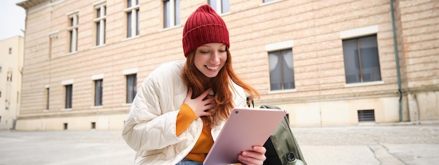 Foto grátis linda mulher ruiva de chapéu vermelho sentada com mochila e garrafa térmica usando tablet digital ao ar livre