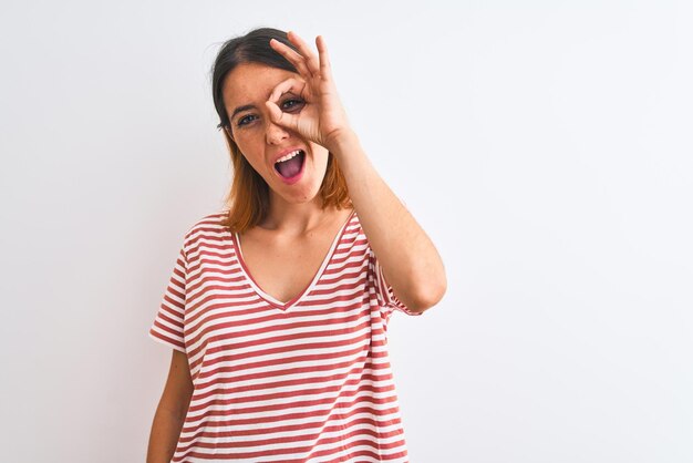 Linda mulher ruiva vestindo camiseta vermelha listrada casual sobre fundo isolado fazendo okey gesto com a mão sorrindo olho olhando por entre os dedos com cara feliz