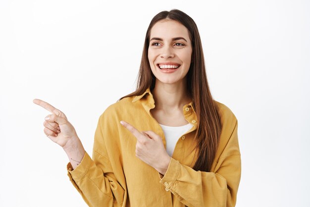 Linda mulher sorridente vira a cabeça e apontando para a esquerda parecendo satisfeita e feliz verificando algo bom mostrando o logotipo do anúncio sobre fundo branco