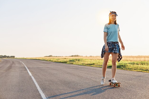 Linda mulher vestindo camiseta, shortinho e faixa de cabelo, andando de skate na rua, olhando ao longe, passando um tempo sozinha com prazer, estilo de vida ativo e saudável.