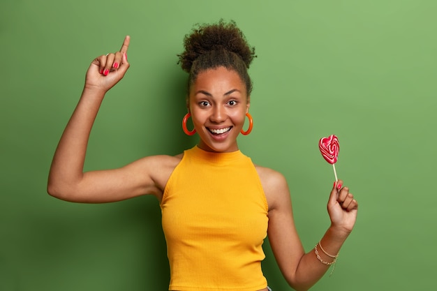 Foto grátis linda namorada despreocupada e atraente dança alegremente com pirulito em forma de coração, se diverte em casa, adora doces e tem bom humor depois de comer doces gostosos, se move com alegria sobre a parede verde vívida