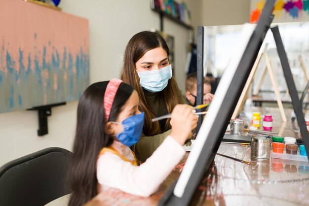 Linda professora de latim e jovem caucasiana usando máscaras faciais estão pintando com um pincel em uma tela em branco para a escola de arte