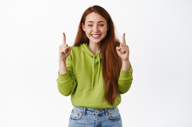 Linda ruiva com sorriso fofo branco, parece feliz e apontando os dedos para cima no anúncio superior, mostra desconto promocional, de pé sobre fundo branco