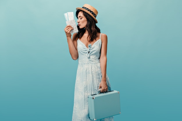 Linda senhora com chapéu de palha tem bolsa moderna e ingressos contra um fundo azul. mulher maravilhosa no vestido leve de verão posando.