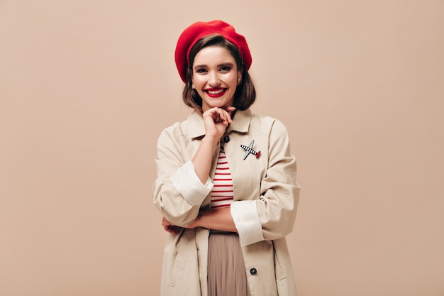 Linda senhora de boina vermelha e sorrisos de trincheira em fundo bege. Mulher jovem feliz com chapéu vermelho e casaco leve está sorrindo para a câmera.