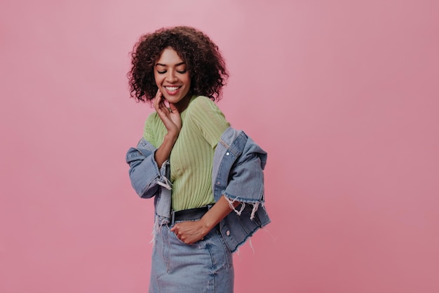 Linda senhora de camiseta verde e jaqueta jeans sorri em fundo rosa Mulher africana morena de saia rindo e posando em pano de fundo isolado