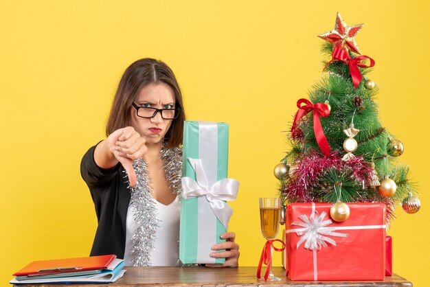 Linda senhora de terno com óculos, mostrando seu dom, fazendo um gesto negativo e sentada à mesa com uma árvore de natal no escritório