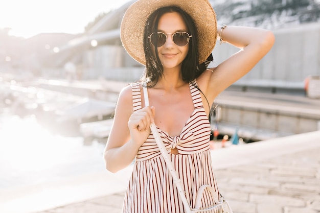 Linda senhora sorridente com cabelo preto, posando com a mão em pé sobre o fundo desfocado. Retrato do close-up de uma jovem satisfeita com a pele bronzeada, passando um tempo na cidade europeia em manhã de verão.