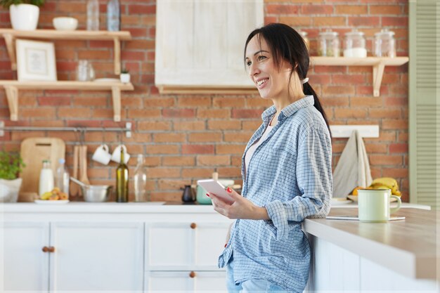 Linda sorridente morena linda mulher usa roupas de casa e fica em pé perto da mesa da cozinha