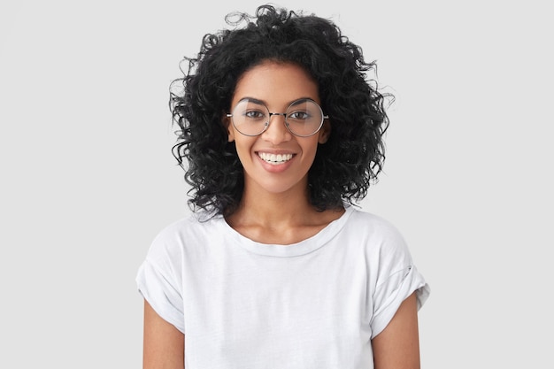 Linda sorridente mulher afro-americana com cabelo crespo, sorriso largo, mostra os dentes brancos, usa camiseta casual e óculos, fica de pé na parede e se alegra com o dia de folga. Mulher jornalista indoor