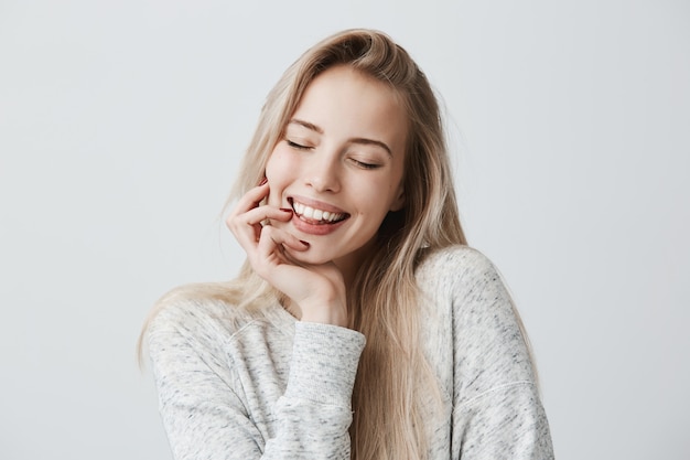 Lindas fêmeas fechando os olhos por causa da alegria, com longos cabelos loiros e um sorriso gentil. alegre mulher segura a mão sob o queixo, agradou a expressão do rosto. expressão facial e emoções positivas