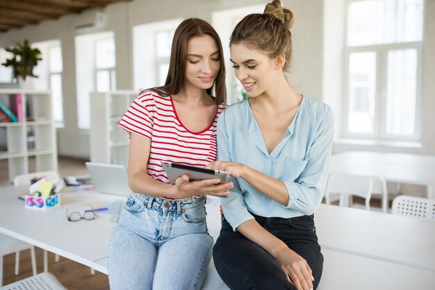 Lindas garotas sorridentes sentadas na mesa sonhadoramente usando o tablet junto com o escritório vazio moderno no fundo