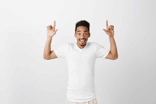 Foto grátis lindo cara de pele escura impressionado e animado apontando os dedos para cima, sorrindo e mostrando promo