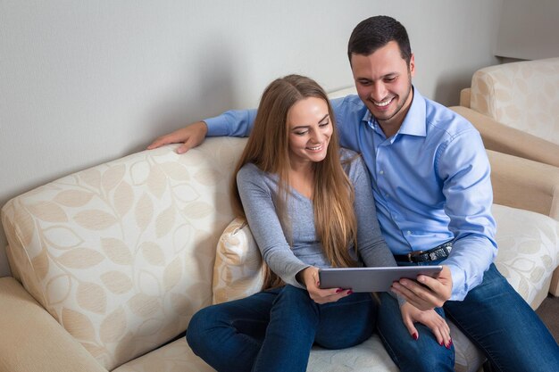 Lindo casal adorável, rindo sentado em um sofá, compartilhando fotos ou outras informações, exibidas em um tablet eletrônico um com o outro