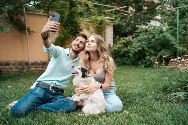 Lindo casal feliz fazendo selfie com seu adorável cachorro no quintal enquanto está sentado na grama