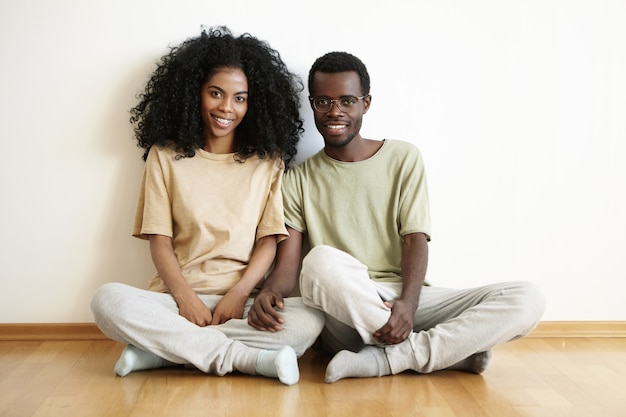 Lindo casal jovem de pele escura casual sentado no chão de madeira depois de se mudar para um novo apartamento