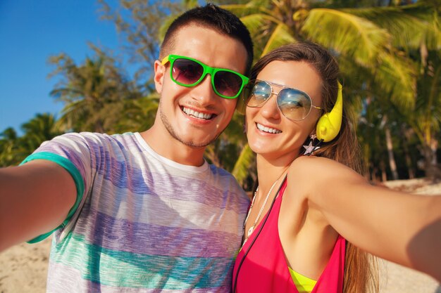 Lindo casal jovem hippie apaixonado fazendo selfie foto em uma praia tropical, férias de verão, felizes juntos, lua de mel, estilo colorido, óculos de sol, fones de ouvido, sorrindo, feliz, se divertindo, positivo