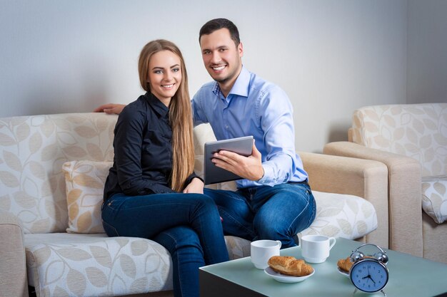 Lindo casal jovem rindo, tomando café com croissants, sentado em um sofá, com tablet eletrônico e olhando para a câmera.