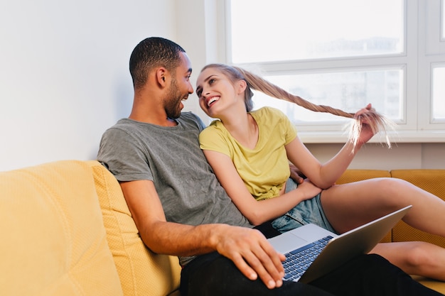 Lindo casal olhando um para o outro e sorrindo. Emparelhe-se sentado no sofá e usando o laptop. Amantes relaxando, curtindo, conversando, mostrando amor. Negro e uma loira juntos em casa.