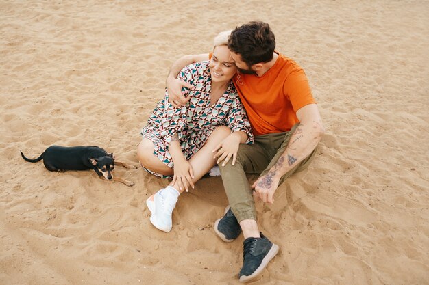 Lindo casal relaxando na areia se abraçando e beijando enquanto brinca com seu cachorro positivo