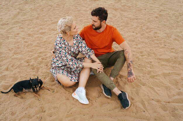 Lindo casal relaxando na areia se abraçando e beijando enquanto brinca com seu cachorro positivo